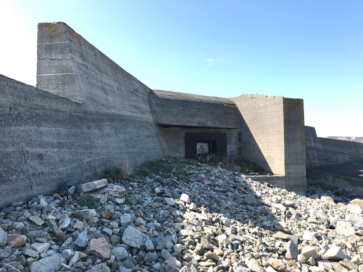 A photo of a bunker on the beach with rocks in the foreground and blue sky in the background.